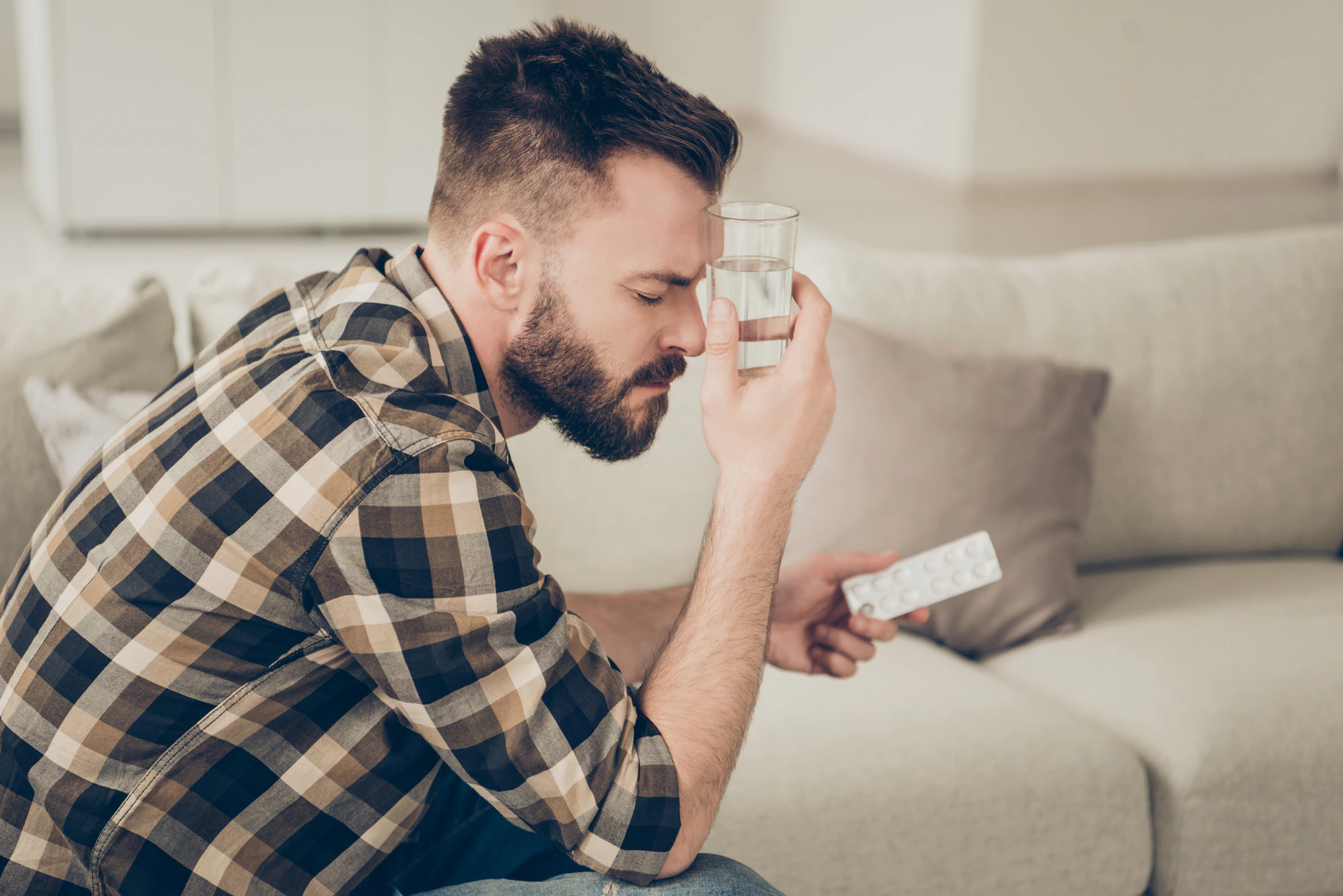 Profile side view photo of concentrated man in brown checkered shirt sitting on the sofa in a bright light room water and tablets in hands leaning a glass against a forehead